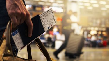 a hand holding passport with a boarding pass tucked into it. A busy airport terminal is in the background. 