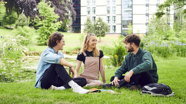 Three students sat on the grass beside a pond. Student flat accommodation is pictured in the background.