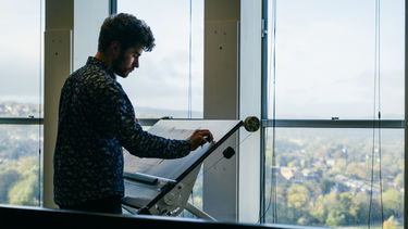 A student stands to work at an upright angled drafting table. Trees and buildings populate the background. 