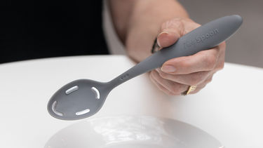 An image of the Tasty Spoon concept, an arm of a woman is holding the spoon above a plate. The spoon is grey.