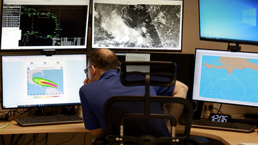 A person sits in front of multiple monitors, with maps of a hurricane 