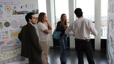 A person with their back to the camera explains their work to three people who are standing beside them.