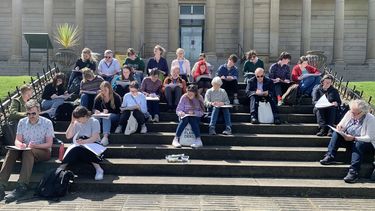 About 20 people sitting on stairs outside a classical looking building. They are all drawing on big pads in the sun