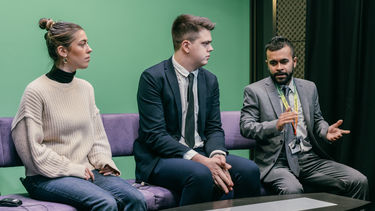 Two students sat on a sofa in front of a green screen. A lecturer is speaking to them. 