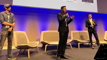 3 men presenting on stage with powerpoint screen on in background