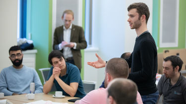 A man in a dark shirts stands in front of peers talking