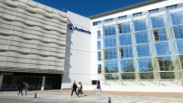 University building with Audencia logo on white wall, one half of building is concrete, the other half windowed. Students walking by on a path in front.