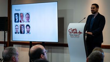 Man with short dark hair and beard, dressed in a dark suit stands at a podium next to a large screen addressing an audience