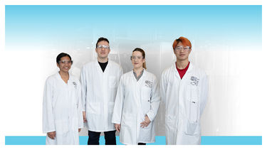 A group shot of four lab technicians in white coats and goggles