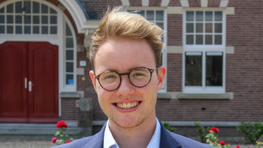 Young man in a suit smiling with a backdrop of an early 20th century house with roses