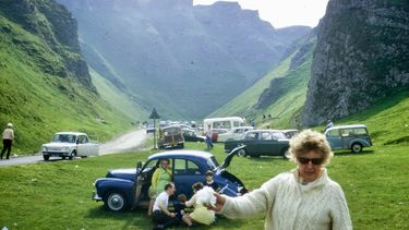 Photograph from the 1970s of people parked in a valley in Castleton in the Peak District - a woman in a cardigan in the foreground in front of a family sitting next to their car