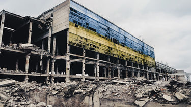 black and white photograph of a bombed building in Ukraine, the only colours being a blue and white flag across the building