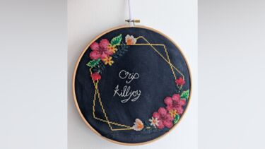 Cross-stiched floral wire wreath on black canvas. In the centre "Crip Killjoy" is stitched in white cursive. At the bottom of the hoop, there is a visible mend where the canvas was too short.