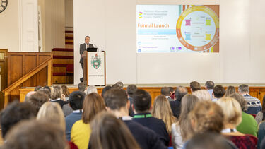 Speaker stood at podium facing a room of people, showing a presentation on a screen