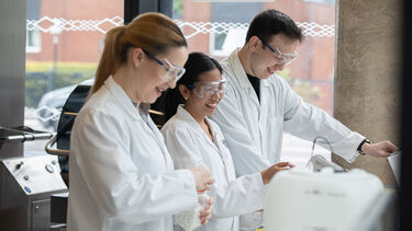 Three smiling students in lab coats and goggles working together in the Diamond powder plant