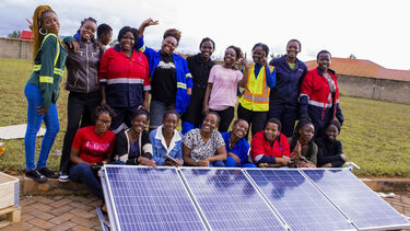 A large group of smiling women stand behind some solar panels