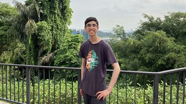Student stood smiling in front of some railings with greenery in the background
