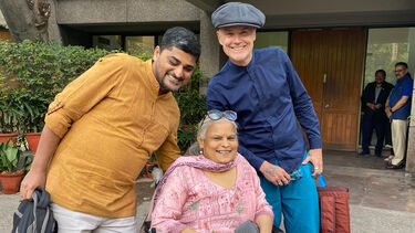 Anita Ghai is seen in her wheelchair, wearing a bright pink dress. On the left is Sandeep Singh wearing an orange top and cream trousers. On the right stands Dan Goodley, wearing a flat cap, blue shirt and trousers. All are smiling together outside a building entrance. 