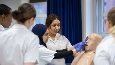 A group of students watch as a nursing student in a blue hijab works on a manikin