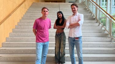 A student stood on a staircase in a University building with two academic members of staff stood either side, all of them looking at the camera.