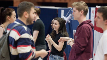 A student stands speaking and smiling with another student at an event. Other people are pictured in the foreground also having a conversation.