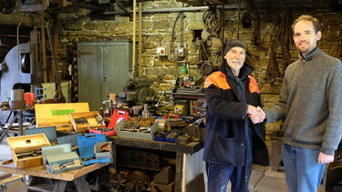 Richard Hodgkinson (University of Sheffield), shaking hands with Ted Young (Wortley Top Forge), standing within the workshop of Wortley Top Forge. Tooling donated to the forge is laid out on benches.