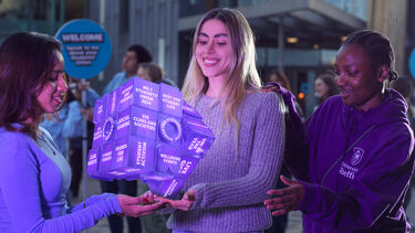 Three University of Sheffield students stood outside the Students' Union smiling while holding a purple fortune telling cube in their outstretched hands. 