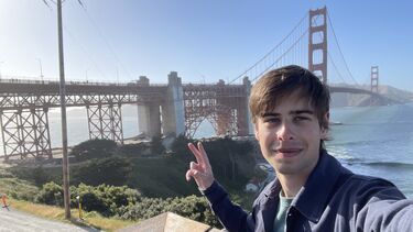 A male student stands outside with the Golden Gate Bridge in the background. He is smiling at the camera and making a peace sign with his hand.