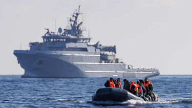 A group of people sit on a dinghy in the water, with a large ship in the distance.
