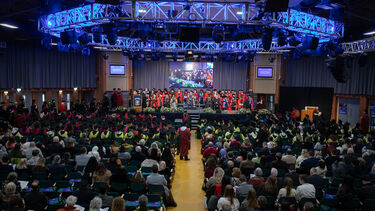 Large group of people wearing graduation robes and caps sit as graduands walk across the centre stage to receive their degree certificate.