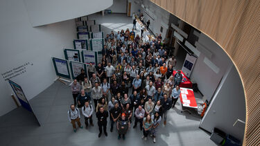 Large group photo from above in a large curved building with white walls and wooden panels.