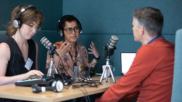 Two women and a man sat in a booth wearing headphones and talking across a table with microphones and recording equipment