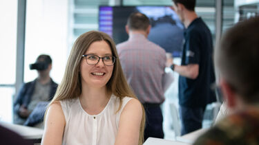 A woman with long hair and glasses smiling with a mad wear a VR headset in the background