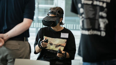  A person sitting down wearing a VR headset and hand held controls with two people in the foreground watching