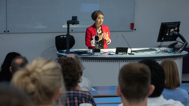 A woman wearing a headscarf and red jacket stands in front of a lectern speaking to the audience in a lecture theatre.