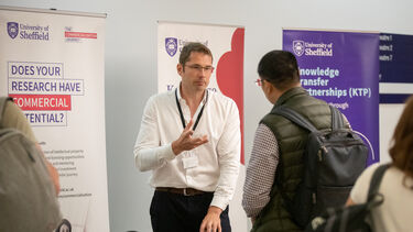 A man with short, light brown hair and glasses wearing a white shirt and black trousers is stood talking to a mat with cropped black hair wearing a checked shirt, green body warmer grey rucksack. In the background are University of Sheffield banners on the tops of research commercial potential and knowledge transfer partnerships.