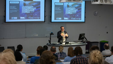 A man wearing a dark grey suit, dark framed glasses and a rainbow lanyard is stood at a lectern speaking to lecture theatre full of people. Behind him are two projector screens.