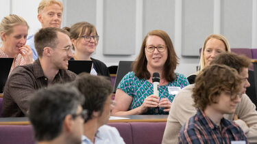 A woman with mid-length straight brown hairs and brown rimmed glasses whearing a green and pink floral dress holds a microphone to ask a question. She's sitting in a lecture surrounded by smiling people.