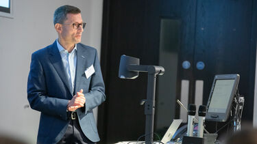 A man with short grey hair and dark rimmed glasses wearing a dark grey suit and pale blue shirt stands in the light in front of a lectern and computer monitor with his hands held together.
