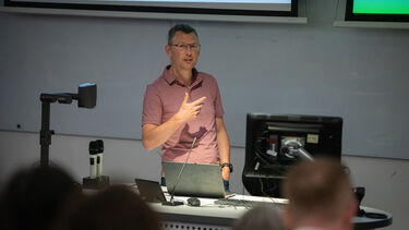 A man with short grey hair and glasses stands, wearing a short-sleeved pink shirt  in front of a lectern speaking 