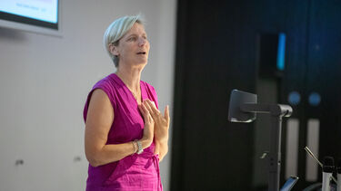 A woman with short grey hair wearing a dark pink dress stand in front of a lectern speaking.