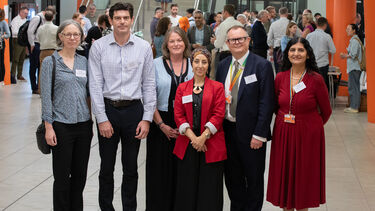 Four women and two men pose for a group photo agains a busy backdrop of people.
