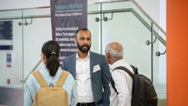 A man with short dark hair and a beard stands in from of a dark Yorkshire MedTech bannerstand talking to a man and a woman wearing rucksacks.