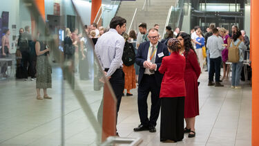 A group of people talking against a busy backdrop of people in a space with a large staircase and bright orange columns.