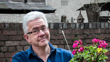 Headshot of a person against a red brick wall outside. They're holding a flower pot and smiling.