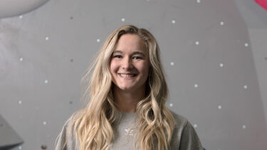 Headshot of a smiling person with long hair in front of an indoor climbing wall