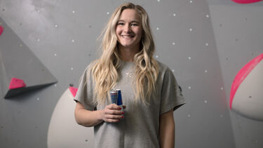 Shauna Coxsey smiling in front of a grey climbing wall, wearing a beige t-shirt, long blonde hair and holding a can of Red Bull