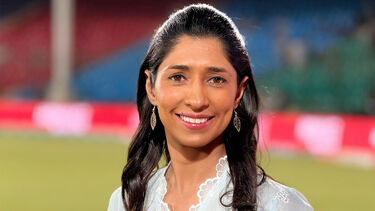 Headshot of a person with long straight hair smiling. Sports stadium pitch/seating is in the background.