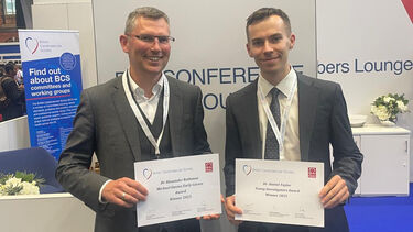 Two smiling men in grey suits holding british cardiovascular society award certificates against a background of conference banners