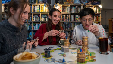 Three people playing a boardgame. There are drinks and snacks on the table.